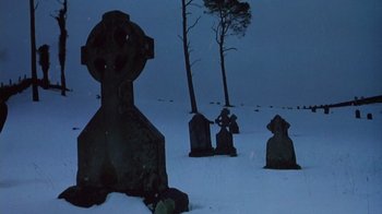 Movie still from “The Dead” (1987), directed by John Huston – A cemetery with a cross and trees in the snow; Wide shot, Low angle