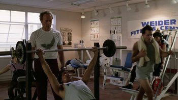 Movie still from “The Dead Pool” (1988), directed by Buddy Van Horn – A man lifting a barbell in a room with other people; Medium shot, Over the shoulder angle