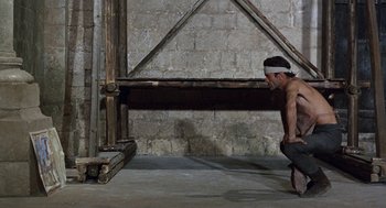 Movie still from “The Decameron” (1971), directed by Pier Paolo Pasolini – A man squatting down in front of a wooden structure; Wide shot, Low angle