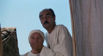 Movie still from “The Decameron” (1971), directed by Pier Paolo Pasolini – A man and a woman standing next to each other with their hands over their mouths; Medium shot, Low angle