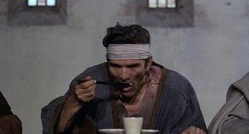 Movie still from “The Decameron” (1971), directed by Pier Paolo Pasolini – A man with a blindfold eating something from a bowl; Medium shot, High angle