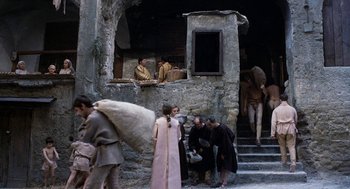 Movie still from “The Decameron” (1971), directed by Pier Paolo Pasolini – A group of people standing outside of a stone building; Wide shot, High angle