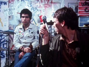 Movie still from “The Decline of Western Civilization” (1981), directed by Penelope Spheeris – A man holding a microphone in front of another man holding a beer; Medium shot, Over the shoulder angle