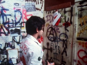 Movie still from “The Decline of Western Civilization” (1981), directed by Penelope Spheeris – A young man standing in front of a wall covered in graffiti; Medium shot, Overhead angle