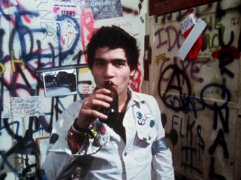 Movie still from “The Decline of Western Civilization” (1981), directed by Penelope Spheeris – A young man drinking a soda in front of a wall covered in graffiti; Medium shot, High angle