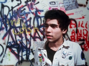 Movie still from “The Decline of Western Civilization” (1981), directed by Penelope Spheeris – A young man standing in front of a wall covered in graffiti; Close Up shot, Overhead angle
