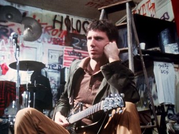 Movie still from “The Decline of Western Civilization” (1981), directed by Penelope Spheeris – A man sitting on a chair holding a guitar in a room; Medium shot, Low angle