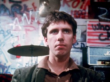 Movie still from “The Decline of Western Civilization” (1981), directed by Penelope Spheeris – A man in a brown jacket is in front of a wall with graffiti on it; Close Up shot, Low angle