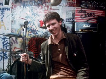 Movie still from “The Decline of Western Civilization” (1981), directed by Penelope Spheeris – A man holding a microphone in front of a microphone stand in a room; Medium shot, Low angle