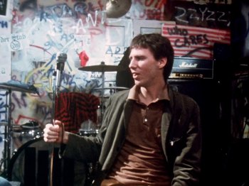 Movie still from “The Decline of Western Civilization” (1981), directed by Penelope Spheeris – A man holding a microphone in front of a wall covered in graffiti; Medium shot, Over the shoulder angle