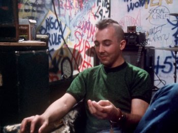 Movie still from “The Decline of Western Civilization” (1981), directed by Penelope Spheeris – A man sitting in front of a wall covered in graffiti; Medium shot, Over the shoulder angle
