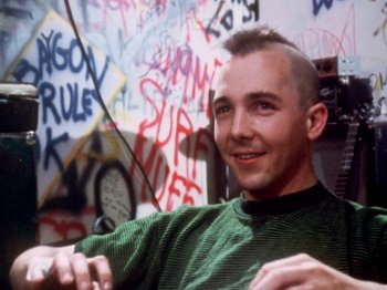 Movie still from “The Decline of Western Civilization” (1981), directed by Penelope Spheeris – A man with a mohawk haircut sitting in front of a wall covered in graffiti; Close Up shot, Over the shoulder angle