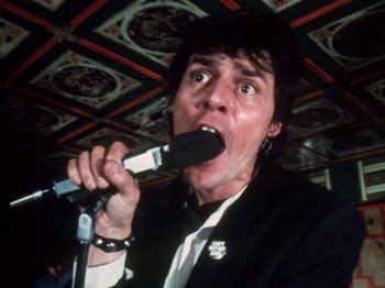 Movie still from “The Decline of Western Civilization” (1981), directed by Penelope Spheeris – A man in a black jacket is singing into a microphone; Close Up shot, Low angle