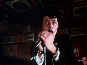Movie still from “The Decline of Western Civilization” (1981), directed by Penelope Spheeris – A man in a black suit is holding a microphone; Medium shot, Low angle