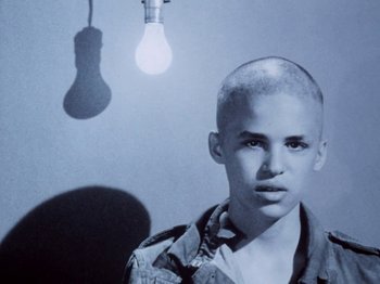 Movie still from “The Decline of Western Civilization” (1981), directed by Penelope Spheeris – A young boy with a shaved head standing in front of a light bulb; Close Up shot, High angle