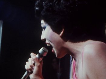 Movie still from “The Decline of Western Civilization” (1981), directed by Penelope Spheeris – A woman is holding a microphone up to her mouth; Close Up shot, High angle