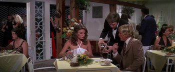 Movie still from “The Deep” (1977), directed by Peter Yates – A woman sitting at a dinner table with two men in the background; Medium shot, Over the shoulder angle