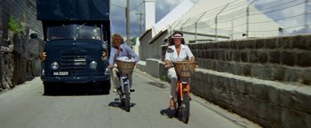 Movie still from “The Deep” (1977), directed by Peter Yates – Two women riding bikes down a street with a bus in the background; Wide shot, Low angle