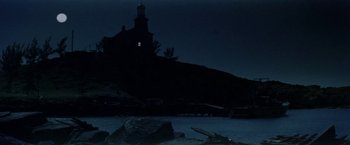 Movie still from “The Deep” (1977), directed by Peter Yates – A lighthouse is lit up at night on the shore of a body of water; Extreme Wide shot, Low angle