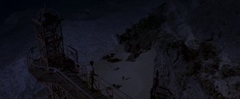 Movie still from “The Deep” (1977), directed by Peter Yates – A man standing on a metal railing next to a body of water at night; Extreme Wide shot, Overhead angle