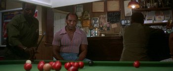 Movie still from “The Deep” (1977), directed by Peter Yates – A man sitting in front of a pool table; Medium shot, Over the shoulder angle
