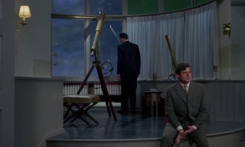 Movie still from “The Devil Rides Out” (1968), directed by Terence Fisher – A man in a suit sitting in front of another man in a suit standing in front of a window; Wide shot, High angle