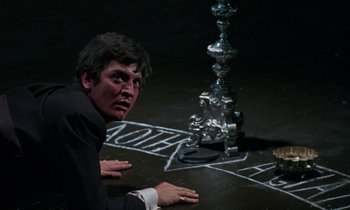 Movie still from “The Devil Rides Out” (1968), directed by Terence Fisher – A man sitting on the ground in front of a silver object; Close Up shot, High angle