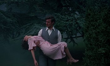 Movie still from “The Devil Rides Out” (1968), directed by Terence Fisher – A man holding a woman upside down in a forest; Medium shot, High angle