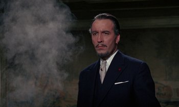 Movie still from “The Devil Rides Out” (1968), directed by Terence Fisher – A man wearing a suit and tie in front of a smoke stack; Medium shot, Low angle