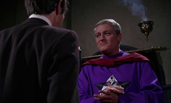 Movie still from “The Devil Rides Out” (1968), directed by Terence Fisher – A man in a purple shirt is talking to another man in a black suit; Medium shot, Over the shoulder angle