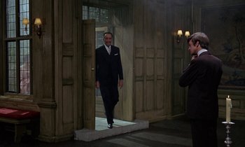 Movie still from “The Devil Rides Out” (1968), directed by Terence Fisher – A man in a suit and tie standing in front of a door; Wide shot, Over the shoulder angle
