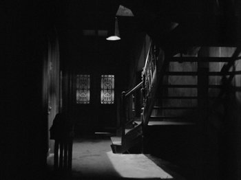 Movie still from “The Devil Strikes at Night” (1957), directed by Robert Siodmak – A black and white photo of a dark room with stairs; Extreme Wide shot, High angle