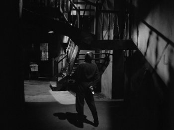 Movie still from “The Devil Strikes at Night” (1957), directed by Robert Siodmak – A man walking in the dark in front of a staircase; Wide shot, High angle
