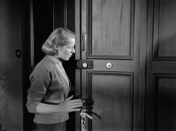Movie still from “The Devil Strikes at Night” (1957), directed by Robert Siodmak – A black and white photo of a woman standing in front of an open door; Medium shot, High angle