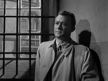 Movie still from “The Devil Strikes at Night” (1957), directed by Robert Siodmak – A black and white photo of a man standing in front of a window; Medium shot, Low angle