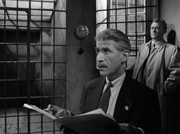 Movie still from “The Devil Strikes at Night” (1957), directed by Robert Siodmak – An older man in a business suit holding papers in front of a door; Medium shot, Over the shoulder angle