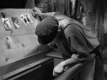 Movie still from “The Devil Strikes at Night” (1957), directed by Robert Siodmak – An old photo of a man working on a piece of wood; Medium shot, High angle