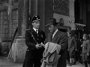 Movie still from “The Devil Strikes at Night” (1957), directed by Robert Siodmak – A man in a suit and hat is talking to a man in a uniform; Medium shot, Low angle