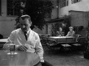 Movie still from “The Devil Strikes at Night” (1957), directed by Robert Siodmak – A man sitting at a table with a cigarette in his mouth; Wide shot, Low angle