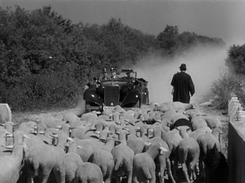 Movie still from “The Devil Strikes at Night” (1957), directed by Robert Siodmak – A herd of sheep walking down a dirt road next to an old car; Wide shot, High angle