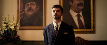 Movie still from “The Devil's Double” (2011), directed by Lee Tamahori – A man in a suit and tie in front of two portraits; Close Up shot, Low angle
