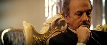 Movie still from “The Devil's Double” (2011), directed by Lee Tamahori – An older man sitting in a chair with a gold frame around his neck; Close Up shot, Low angle
