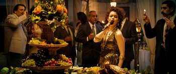 Movie still from “The Devil's Double” (2011), directed by Lee Tamahori – A woman in a gold dress singing into a microphone at an event; Medium shot, High angle