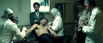 Movie still from “The Devil's Double” (2011), directed by Lee Tamahori – A man getting a tattoo on his chest while another man looks on; Medium shot, Low angle