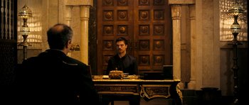 Movie still from “The Devil's Double” (2011), directed by Lee Tamahori – A man sitting at a desk in front of a wooden door; Wide shot, Over the shoulder angle