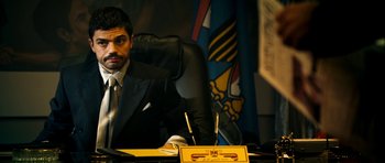 Movie still from “The Devil's Double” (2011), directed by Lee Tamahori – A man sitting at a desk in front of a flag; Medium shot, Over the shoulder angle