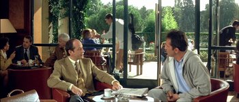 Movie still from “Le Dîner de Cons” (1998), directed by Francis Veber – Two men sitting at a table in front of a glass door; Medium shot, Over the shoulder angle