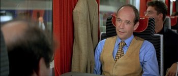 Movie still from “Le Dîner de Cons” (1998), directed by Francis Veber – A man sitting on top of a train next to a red curtain; Medium shot, Over the shoulder angle