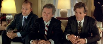 Movie still from “Le Dîner de Cons” (1998), directed by Francis Veber – A man in a suit sitting in front of two other men; Medium shot, Low angle
