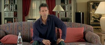 Movie still from “Le Dîner de Cons” (1998), directed by Francis Veber – A man sitting on top of a couch in a living room; Medium shot, Over the shoulder angle